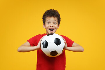 Cute little soccer player boy wearing basic red t shirt, holding soccer ball while looking at camera with amazed face expresion, isolated veryellow background