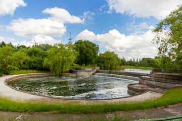 A small lake located in a park in Europe. Natural shoreline covered with grass. Calm water surface with reflective sky in summer.