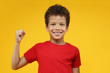 Portrait of happy smiling beautiful African American boy posing posing in studio making winner's gesture clenching his fist, isolated over bright colored orange yellow background