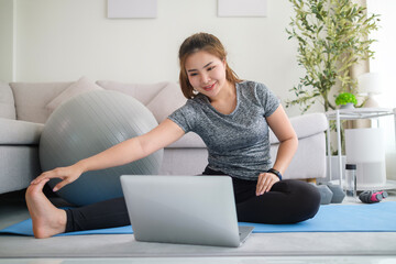 Smiling young sporty woman doing fitness exercise on mat and watching online tutorial on laptop at...