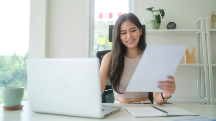 Attractive young businesswoman holding document and working with laptop at office.
