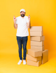 Full-length shot of delivery man among boxes over isolated yellow background with fingers crossing and wishing the best