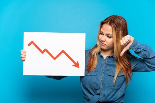Teenager girl over isolated blue background holding a sign with a decreasing statistics arrow symbol and doing bad signal