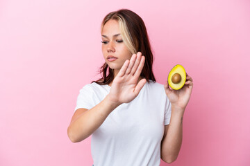 Young Russian woman holding avocado isolated on pink background making stop gesture and disappointed