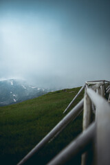Beautiful dramatic scenery on top of a hill with snowy background mountian covered in fog and clouds seen from a path with wooden fence and vibrant green grass.
