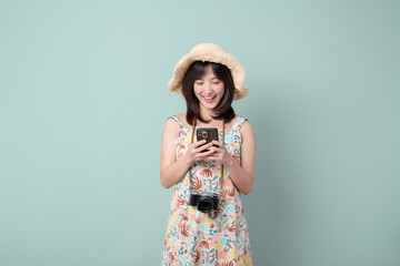 Travel and vacation concept. Happy Asian woman wearing casual dress and hat with camera while using mobile phone on vacation or travel theme isolated on pastel green background.