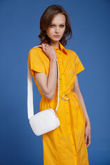 High fashion photo of a beautiful skinny young woman in pretty long yellow dress with short sleeves, white handbag posing on blue background. Slim figure.