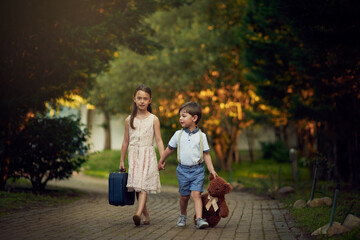 Holding hands, suitcase and teddy bear with children walking on path in forest for travel to school. Destination, evening or luggage with lost brother and sister kids outdoor in park or woods