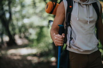 Hiker wearing a sleeveless hoodie and backpack, holding a trekking pole while walking through a forest trail, capturing the essence of outdoor adventure and exploration.