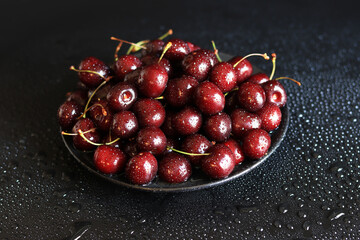 Cherries in a black plate on a dark wet background, close-up. Wet sweet ripe cherries. Dark red cherries with green stems covered with fine splashes of water. Summer berries, vitamins