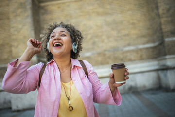 Fototapeta premium African American older woman wearing headphones in the city center. Wearing headphones and drinking coffee.