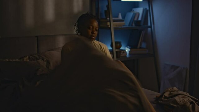 Medium shot of young African American lady getting up from bed at night due to insomnia, experiencing intrusive thoughts and flashbacks to traumatic situations in bedroom