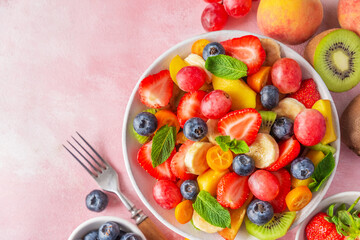 Summer fresh fruit salad in a bowl with fork on pink background. Top view. Healthy food for breakfast. Mixed strawberries, grapes, banana, kiwi, blueberries, peach, kumquat and mint for diet lunch