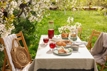 Stylish table setting with beautiful spring flowers, fruit drink and pie in garden