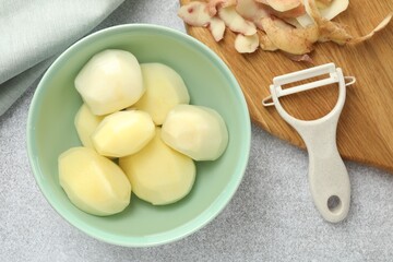 Fresh raw potatoes in bowl, peeler and peels on light grey table, top view