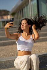 Chica joven en verano sonriendo haciendo fotos con el móvil en un lugar de mar y bonito. Pelo rizado y brillante. 