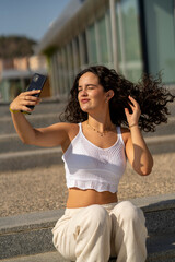 Chica joven en verano sonriendo haciendo fotos con el móvil en un lugar de mar y bonito. Pelo rizado y brillante. 