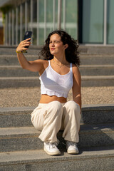 Chica joven en verano sonriendo haciendo fotos con el móvil en un lugar de mar y bonito. Pelo rizado y brillante. 