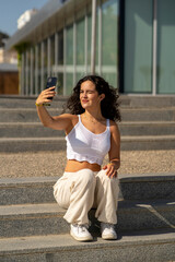 Chica joven en verano sonriendo haciendo fotos con el móvil en un lugar de mar y bonito. Pelo rizado y brillante. 