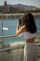 Chica joven en verano sonriendo haciendo fotos con el móvil en un lugar de mar y bonito. Pelo rizado y brillante. 