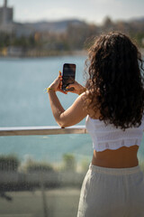 Chica joven en verano sonriendo haciendo fotos con el móvil en un lugar de mar y bonito. Pelo rizado y brillante. 