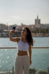 Chica joven en verano sonriendo haciendo fotos con el móvil en un lugar de mar y bonito. Pelo rizado y brillante. 