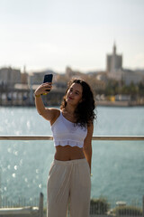 Chica joven en verano sonriendo haciendo fotos con el móvil en un lugar de mar y bonito. Pelo rizado y brillante. 