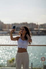 Chica joven en verano sonriendo haciendo fotos con el móvil en un lugar de mar y bonito. Pelo rizado y brillante. 
