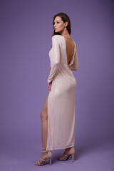 High fashion photo of a beautiful elegant young woman in pretty cream long dress with sleeves, cutout on the leg posing on purple, lilac background. Slim figure. 