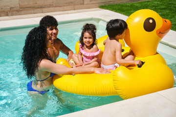 A family of four is enjoying a day at the pool on a yellow inflatable duck. Scene is lighthearted and fun