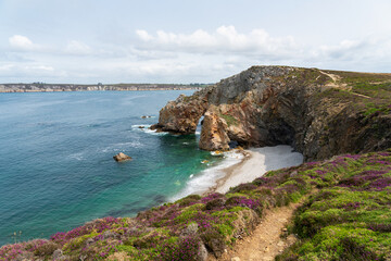 Panorama de l'anse de Dinan, sous un ciel nuageux, avec les eaux turquoises de la mer d'Iroise en toile de fond et un parterre de bruyères fleuries.