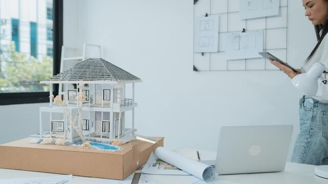 Beautiful caucasian architect focus on using tablet to check house model on meeting table with blueprint, house model and architectural document scatter around at meeting room. Immaculate.