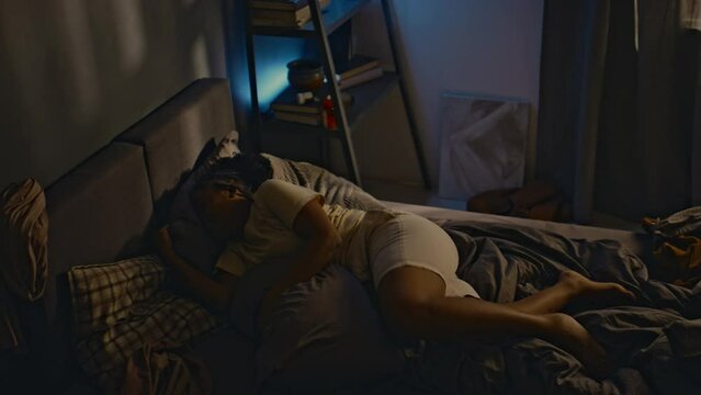 High angle side shot of young Black woman with ptsd rolling over in bed, waking up from terrifying nightmares and sitting up at night, living with traumatic memories