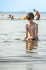 A young boy stands in shallow water at the beach, focused on the water in his hands. In the background, several people enjoy the water on a sunny day.