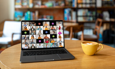 A laptop computer sits on a wooden table in a cafe. The laptop screen displays a video conference...