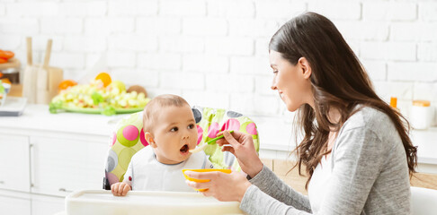 A young mother is feeding her baby in a high chair in the kitchen. The baby is sitting in a brightly colored high chair and is looking at the mother with a wide-eyed expression, copy space