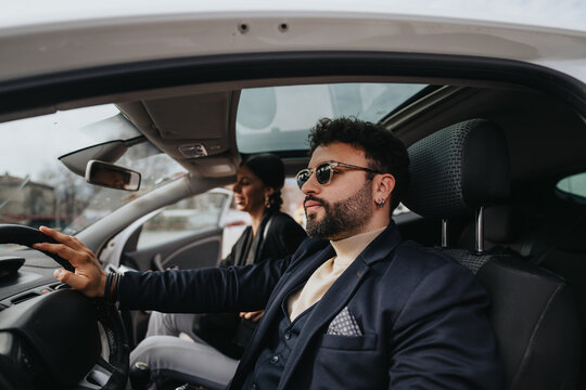 A well-dressed man in sunglasses driving a car, with a focused expression and a woman in the passenger seat, possibly commuting or traveling together in an urban setting.