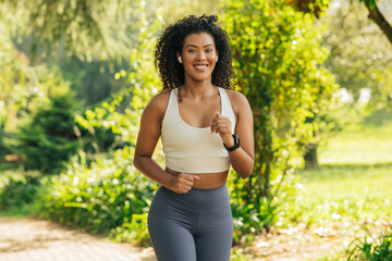 A woman wearing a white sports bra and gray leggings runs on a path through a park. She is smiling...