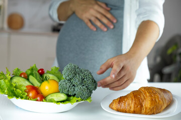 Pregnant woman choose croissant or fresh vegetables think about what to eat. The concept of healthy and unhealthy food, mindful eating during pregnancy. Diet while gestational diabetes.