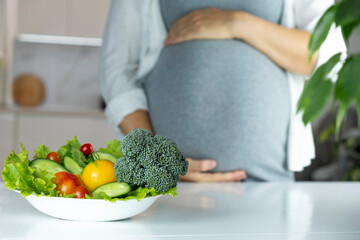 Pregnant woman with bowl of fresh vegetables, broccoli, leaf salad, tomato on table. Healthy nutrition and diet during pregnancy. Prenatal care, weight control, happy motherhood, new life.