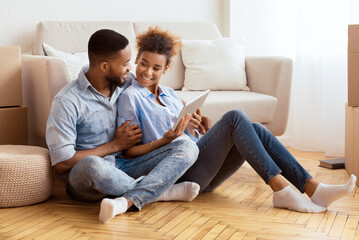 Young Black Couple Using Digital Tablet Sitting On Floor Among Moving Boxes Relaxing In New Apartment. House Ownership