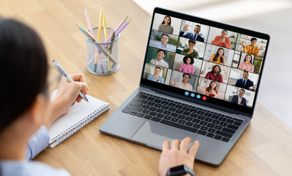 Lady woman sits at a desk, taking notes with a pen and notepad as she participates in a virtual meeting on her laptop. The screen displays a grid of video calls