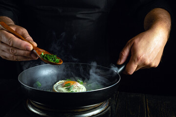The chef cooks a delicious fried eggs in a frying pan. Adding herbs with a spoon to the hot fried egg.