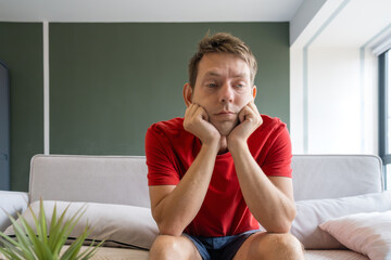 Bored sad man in a red shirt sits on couch at home with his chin resting on his hands. Tired...