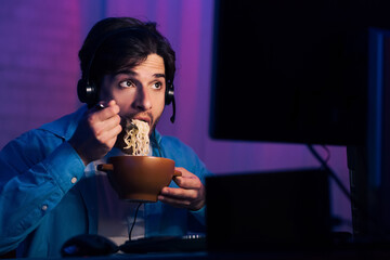 A young man wearing a headset sits in front of a computer monitor in a dimly lit room. He is holding a bowl of instant noodles and eating with chopsticks while looking at the computer screen.