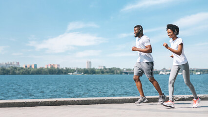 Fototapeta premium Morning jog. Young afro couple running along river embankment in city. Copy space, panorama