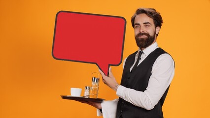 Classy waiter holds speech bubble icon on camera, working to create new advertisement using empty red cardboard. Experienced employee in suit presenting blank carton billboard, serving personnel.