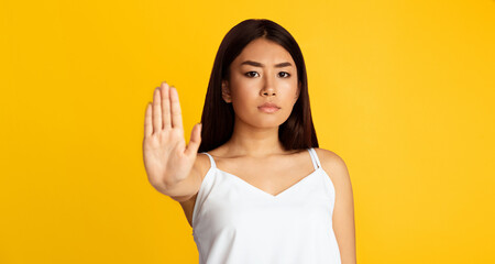 A young Asian woman stands against a bright yellow background. She is facing the camera and holding out her left hand with her palm facing forward in a stop gesture