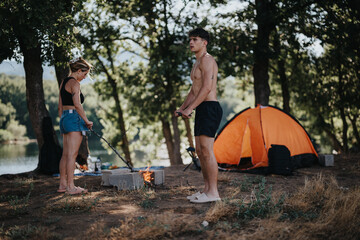 A young couple setting up a barbeque fire at a beautiful mountain picnic spot with an orange camping tent in the background.