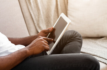 A man is sitting on a couch indoors, using a tablet with his right hand. He is wearing a white t-shirt and jeans. The image is taken from a close-up perspective, focusing on his hands